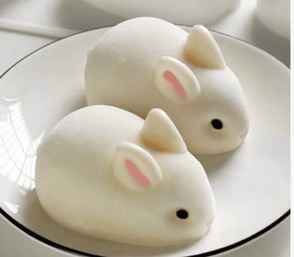After a delightful Saturday shopping, enjoy two cute, white bunny-shaped desserts on a round black-rimmed plate. Each bunny features small pink ears and black eyes, resembling marshmallow or mochi treats that are almost too adorable to eat.